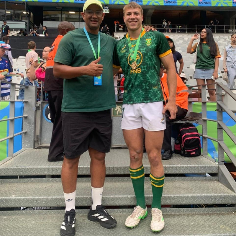 A South Africa sevens player wearing a gold medal, photographed with a supporter in the stands